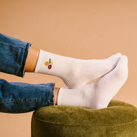 A person wearing The Darling Effect Fruit Market Embroidered Crew Socks, featuring colorful fruit designs, relaxes on a green ottoman. They are in blue jeans with a beige background.