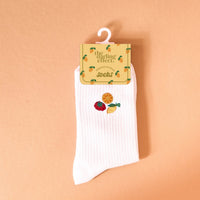 The Darling Effect Fruit Market Embroidered Crew Socks: white women’s socks with a ribbed texture, featuring colorful embroidered strawberry, orange slice, and lemon wedge designs, shown against a peach background.