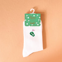 The Darling Effect Golf Embroidered Crew Socks, featuring a white ribbed design with a small green golf course and pink flag, are shown hanging from green packaging against a peach background.