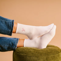 The Darling Effect Pink Bow Embroidered Crew Socks, featuring a small pink bow on the ankle, are styled with blue jeans and shown resting on a green ottoman against a tan background.