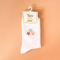 The Darling Effect’s Seashells Embroidered Crew Socks feature white ribbed fabric with colorful seashell and starfish details, displayed on a retail hook against a peach backdrop for a playful finishing touch.