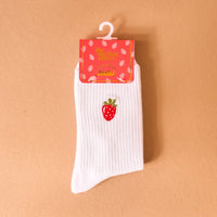 A single white Strawberry Embroidered Crew Sock by The Darling Effect, featuring a red strawberry patch and ribbed design, is shown on a peach background with a strawberry-illustrated label and plastic hanger.