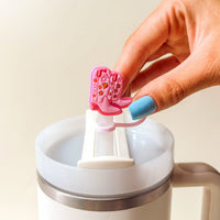 A hand with blue-painted nails holds The Darling Effect's Classic Cowgirl Straw Topper Set—a pink cowboy boot-shaped silicone accessory attached to a tumbler straw—perfect for adding fun and style to your drinkware collection.