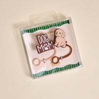 The Darling Effect Dog Mom Straw Topper Set includes two silicone toppers: one with "DOG MOM" and a paw print, and one shaped like a smiling cartoon dog. Both come on a striped green and white background.