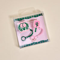 The Darling Effect Pickleball Social Club Straw Topper Set includes two silicone toppers: one green with rackets and “PICKLEBALL SOCIAL CLUB,” and one pink with a paddle and tree—fun drinkware accessories on a cream background.