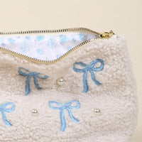 The Bows Blue Teddy Pouch by the-darling-effect features cream sherpa fabric, blue embroidered bows, pearl accents, a gold zipper, and a white floral lining—ideal for mixing and matching with your favorite teddy pouches or accessories.