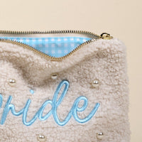 The Bride Teddy Pouch by the-darling-effect is a cream sherpa pouch with pearl accents, light blue embroidered text, a gold zipper, and a blue-and-white checkered lining—a chic accessory for any occasion.