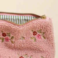A close-up of the-darling-effect's Dainty Floral Pink Teddy Pouch, featuring pink sherpa with embroidered red and pink flowers, a gold zipper, and a green and white striped fabric lining inside.