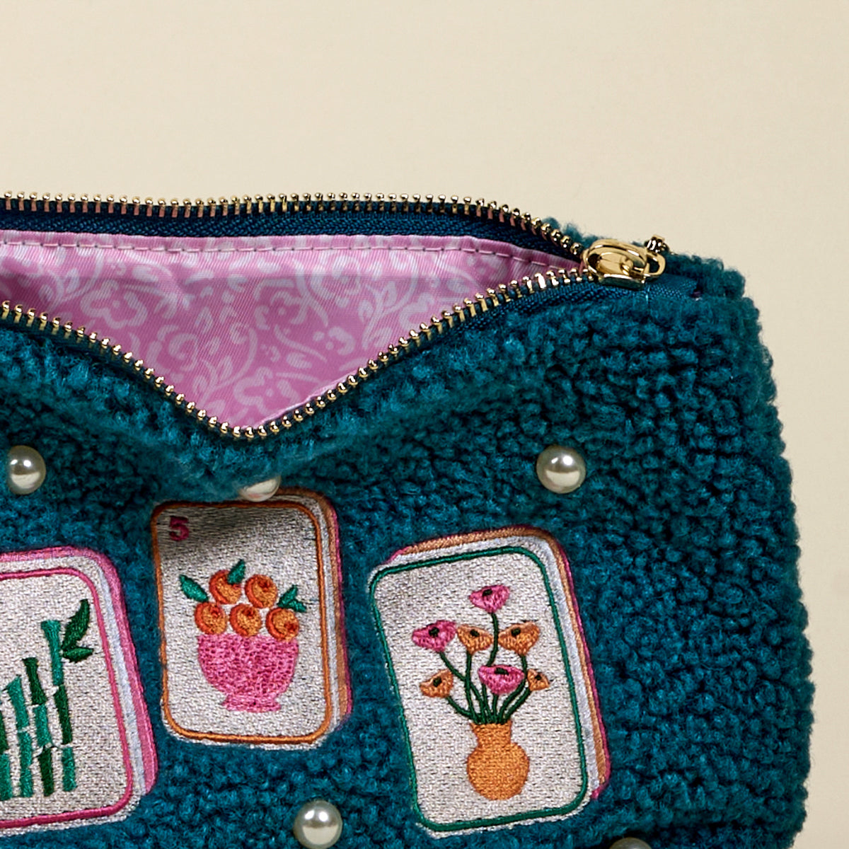 A close-up of The Darling Effect’s "It's Mahj Time Teddy Pouch" in textured teal with a gold zipper, featuring embroidered bamboo, oranges, flowers, decorative pearls, and a pink patterned lining—perfect for your accessory set.