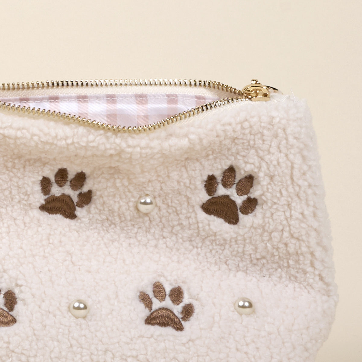 The Darling Effect’s Paws & Pearls Teddy Pouch features a cream sherpa finish with brown paw prints, round pearl-like accents, a gold zipper, and pink and white striped lining—an adorable addition to your teddy pouch collection.