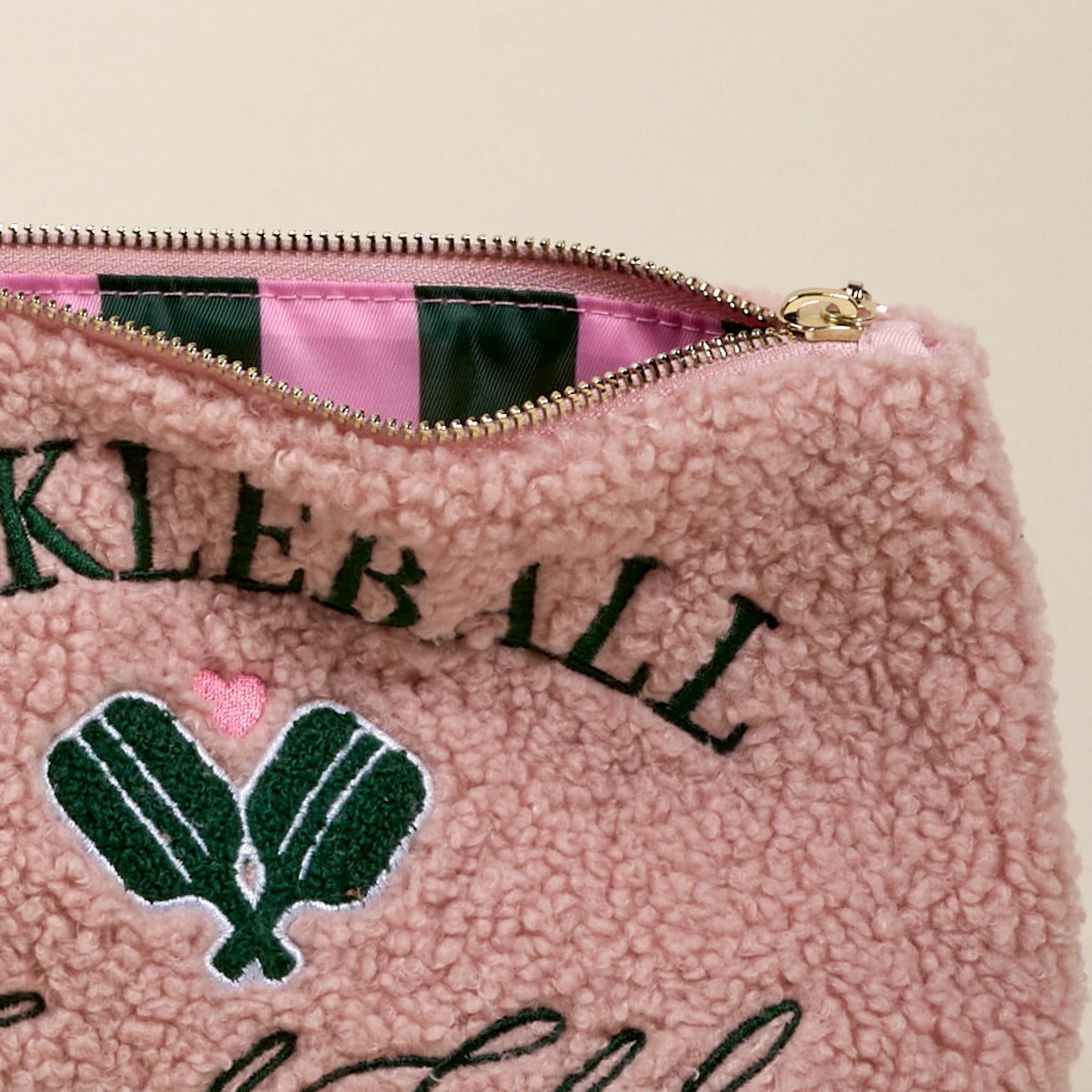 A close-up of The Darling Effect Pickleball Social Club Teddy Pouch shows pink sherpa fabric, green embroidered pickleball paddles, a pink heart, and open zipper revealing a pink and green striped interior—perfect for any accessory set.