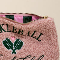 A close-up of The Darling Effect Pickleball Social Club Teddy Pouch shows pink sherpa fabric, green embroidered pickleball paddles, a pink heart, and open zipper revealing a pink and green striped interior—perfect for any accessory set.