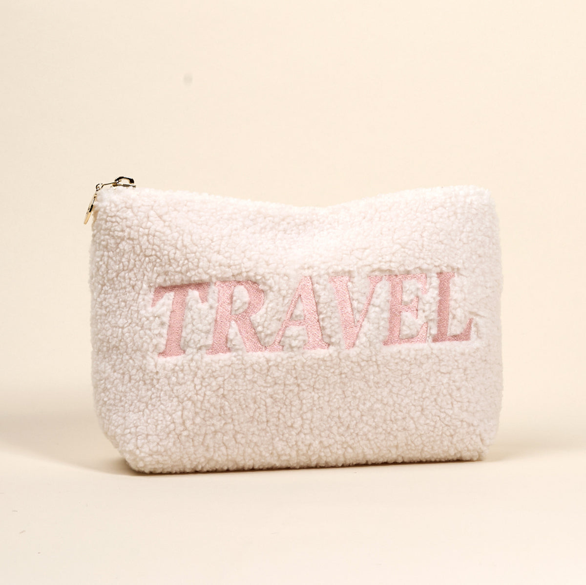The Darling Effect’s Travel Edit Teddy Pouch is a fluffy white sherpa zippered pouch that stands upright, with “TRAVEL” boldly embroidered in large pink letters on the front.