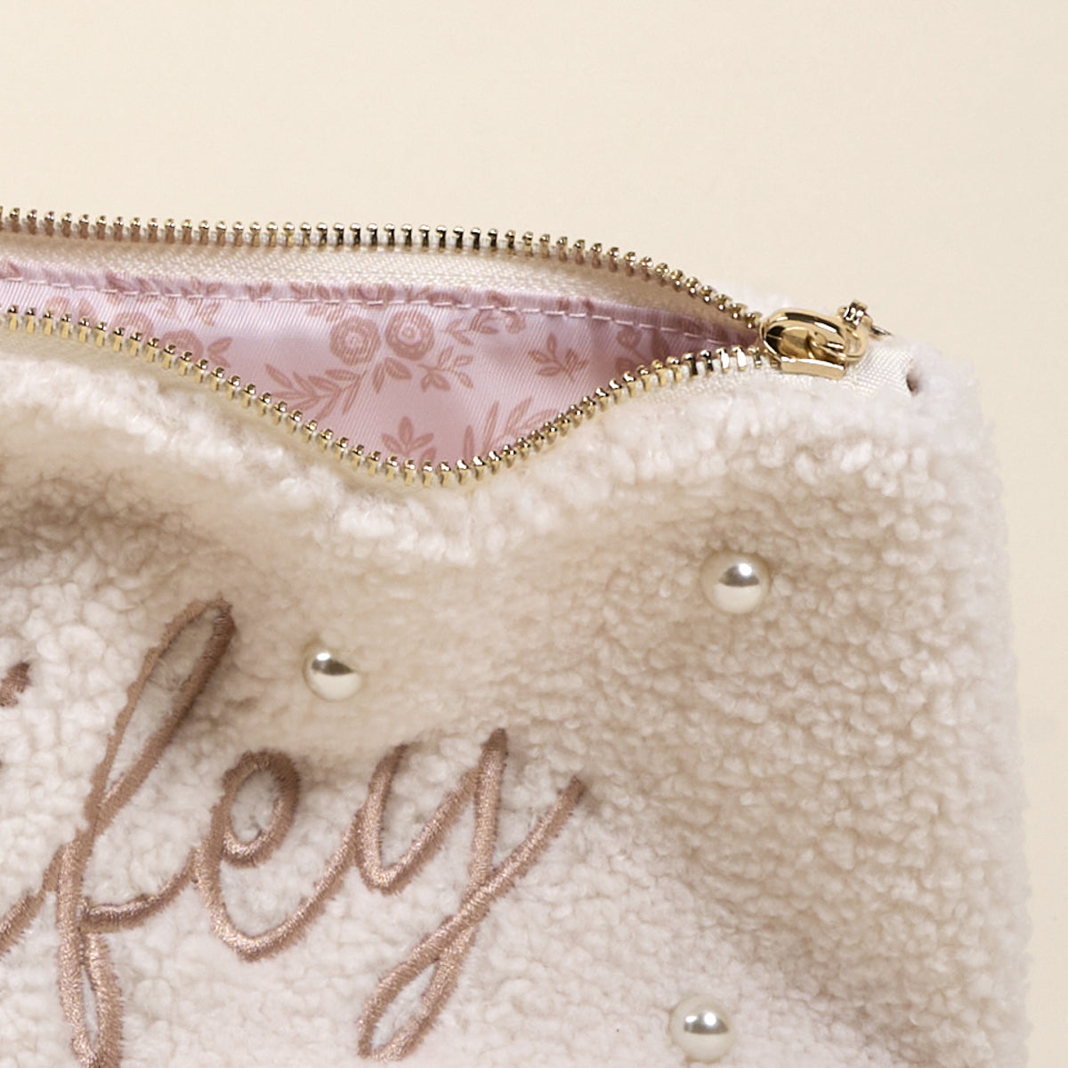 The Darling Effect’s Wifey & Pearls Teddy Pouch features soft cream sherpa, decorative pearls, light brown “Wifey” embroidery, a gold zipper, and a pink floral inner lining.