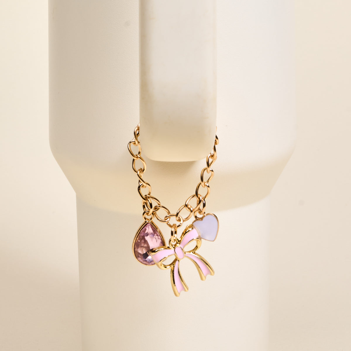 The Darling Effect’s Blushing Beauty Tumbler Charm, featuring a gold chain with a pink teardrop gem and pink-purple enamel bow, is shown hanging on a white cylindrical stand against a beige background.