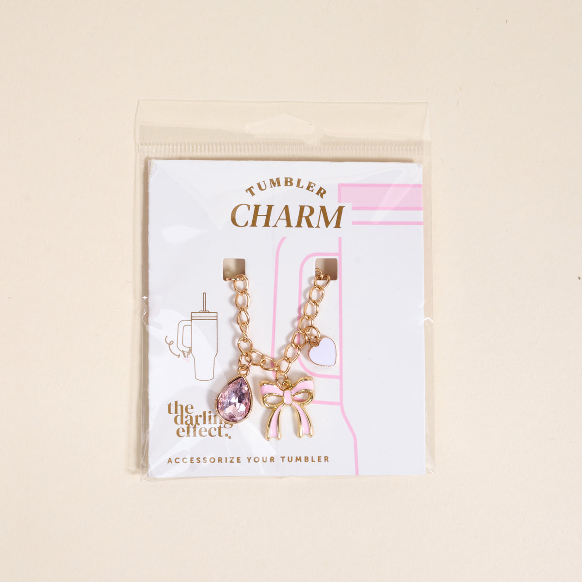The Darling Effect’s Blushing Beauty Tumbler Charm set includes a gold chain with pink gem, bow, and heart charms, displayed on a white and pink card labeled “Tumbler Charm” and “ACCESSORIZE YOUR TUMBLER.”.