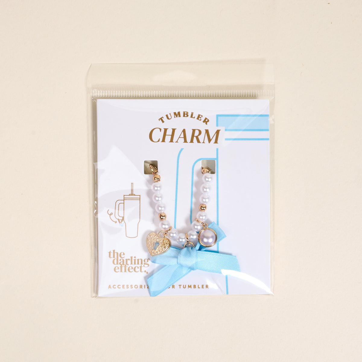 The Darling Effect's Daydreamer Tumbler Charm features white and gold beads, a gold heart, pearl, blue ribbon, and comes in a clear bag with a card labeled “Tumbler Charm” and “The Darling Effect.”.