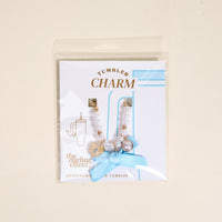 The Darling Effect's Daydreamer Tumbler Charm features white and gold beads, a gold heart, pearl, blue ribbon, and comes in a clear bag with a card labeled “Tumbler Charm” and “The Darling Effect.”.
