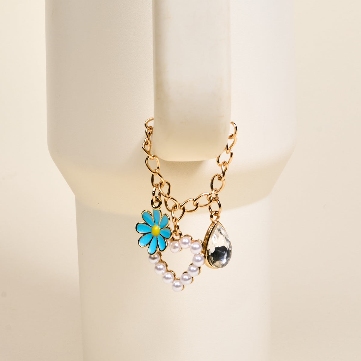 The Darling Effect's Fairytale Tumbler Charm, a gold chain bracelet with a blue enamel daisy, pearl heart outline, and teardrop crystal, is shown on a cream mannequin wrist—perfect for pairing with your favorite tumbler accessory.