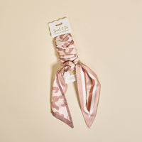 The Darling Effect’s Born Wild Twirl & Tie Scarf Accessory is a soft pink and beige animal print scrunchie with an attached ribbon, shown on cream with a “twirl & tie” label—perfect for unlimited styling.
