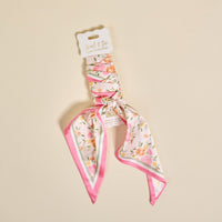The Darling Effect's Endless Daydream Cream Twirl & Tie Scarf Accessory features a pink and white floral scrunchie with a versatile scarf detail, shown on a cream background. The tag reads "Twirl & Tie Scarf Accessory." Ideal for endless styling options.