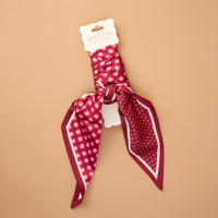 A Gingham Burgundy Twirl & Tie Scarf Accessory by The Darling Effect featuring a maroon, pink, and white satin design with checks and polka dots is neatly tied and attached to a product tag labeled "twist & tie great accessory," displayed against a tan background. This versatile scarf offers endless styling options to complement any outfit.