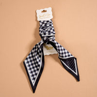 The Darling Effect’s Gingham Black Twirl & Tie Scarf Accessory is shown on a beige background, featuring a classic black and white check pattern and offering versatile styling options for any outfit.