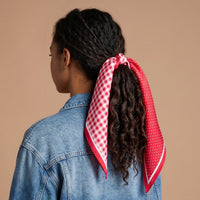 A person with curly hair, seen from behind, wears a denim jacket and has The Darling Effect’s Gingham Red Twirl & Tie Scarf Accessory—featuring white polka dots and a classic gingham pattern—tied around their ponytail, set against a plain beige background.