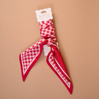 The Gingham Red Twirl & Tie Scarf Accessory by The Darling Effect features red satin adorned with white checkered and polka dot patterns, elegantly tied around a white card on a light brown background, providing versatile styling options for any look.