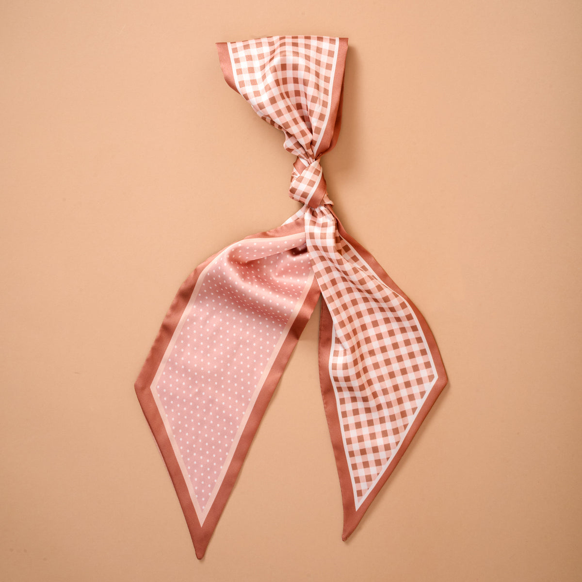 The Darling Effect's Gingham Tan Twirl & Tie Scarf Accessory is showcased on a peach-colored background, featuring one side with gingham checks and the other with playful polka dots—ideal for versatile styling when tied in a loose knot.