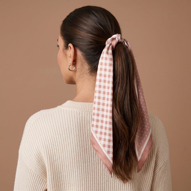 A woman with long brown hair in a low ponytail, secured with The Darling Effect’s Gingham Tan Twirl & Tie Scarf Accessory, wears a cream ribbed sweater and is shown from behind against a beige background.