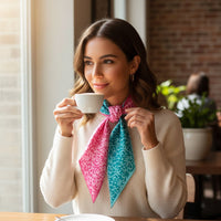 A woman with wavy brown hair in a cream sweater smiles thoughtfully at a café table, holding a white cup as sunlight streams in. She wears The Darling Effect's Pattern Play Twirl & Tie Scarf Accessory in pink and teal.