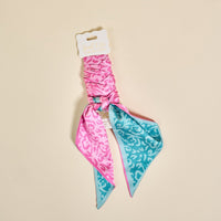 The Darling Effect’s Pattern Play Twirl & Tie Scarf Accessory features a reversible pink and turquoise pattern, tied at the center and displayed on a “twirl & tie scarf accessory” card, adding chic versatility to any outfit.