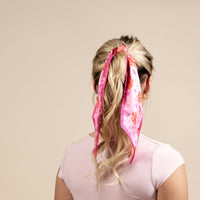 A woman with blonde hair in a ponytail wears a light pink shirt, styled with The Darling Effect’s Poppin' Petals Hot Pink Twirl & Tie Scarf Accessory, adding boutique charm against a plain beige background.