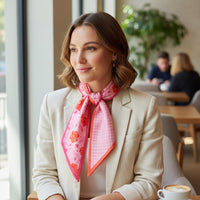A woman with shoulder-length brown hair, wearing a cream blazer and The Darling Effect's Poppin' Petals Hot Pink Twirl & Tie Scarf Accessory, sits at a bright cafe table with coffee; two people blurred in the background enhance the boutique vibe.