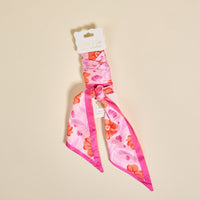 The Darling Effect's Poppin' Petals Hot Pink Twirl & Tie Scarf Accessory, featuring orange and red flowers, comes knotted with a white tag—versatile for styling in hair or as a bag accent against a light beige backdrop.
