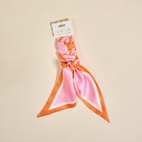 The Darling Effect's Sicily Stripes Pink Twirl & Tie Scarf Accessory features pink and orange stripes, a white tag with "twirl & tie," and offers versatile styling options for any outfit.