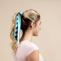 A woman with blonde hair in a ponytail, tied with The Darling Effect's Sicily Stripes Teal Twirl & Tie Scarf Accessory, wears a light pink shirt and gold hoop earrings, facing sideways against a plain beige background.