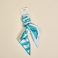 The Darling Effect's Sicily Stripes Teal Twirl & Tie Scarf Accessory, featuring teal and white stripes, is neatly tied at the center and displayed on a branded card against a beige background—perfect for versatile outfit styling.