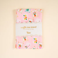 A pink decorative tea towel with framed flower and bird prints, folded and wrapped with a label that reads "It’s Mahj Time Waffle Tea Towel" from The Darling Effect. Absorbent and perfect for everyday kitchen use.