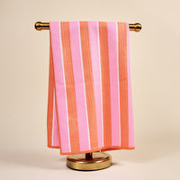 The Darling Effect’s Sicily Stripes Pink Waffle Tea Towel, featuring pink and orange stripes and a textured waffle pattern, is displayed neatly on a gold towel stand against a plain beige background.