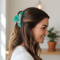 A woman with long, wavy brown hair smiles in profile, wearing The Darling Effect's Mahj Word Up Claw Clip in green. A potted plant and a hanging light are softly blurred in the background.