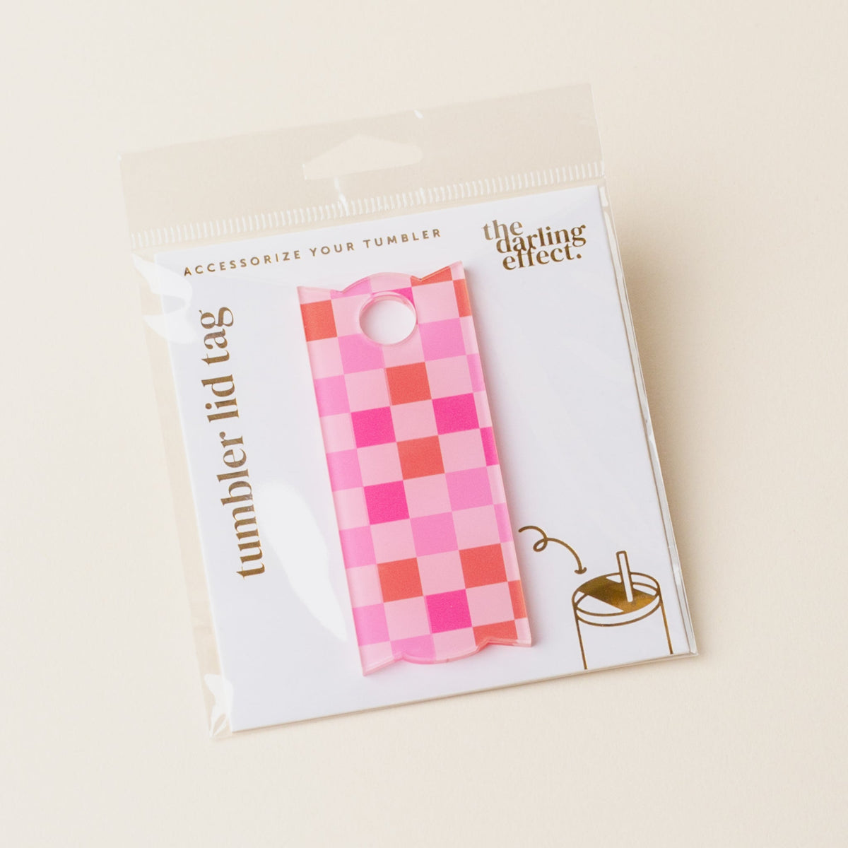 A Hot Pink Checker Tumbler Lid Tag by The Darling Effect is shown in its packaging. An illustration demonstrates how to attach the tag to a Stanley Quencher 2.0, with text reading “tumbler lid tag.”.