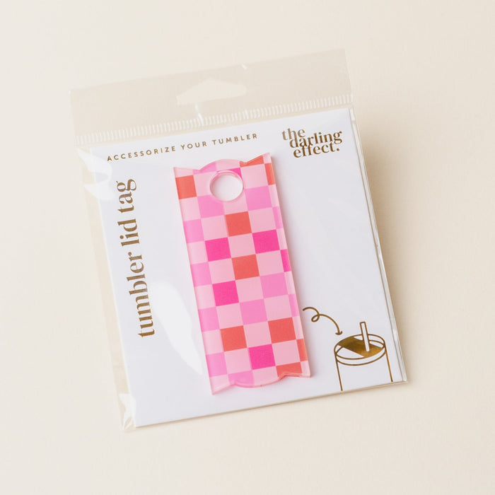 A Hot Pink Checker Tumbler Lid Tag by The Darling Effect is shown in its packaging. An illustration demonstrates how to attach the tag to a Stanley Quencher 2.0, with text reading “tumbler lid tag.”.
