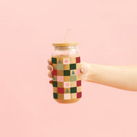 A hand holds The Darling Effect’s Round Glass Cup - Holiday Check, which features a bamboo lid and straw and is decorated with a pink, green, and yellow patchwork pattern with trees and flowers, set against a light pink background.