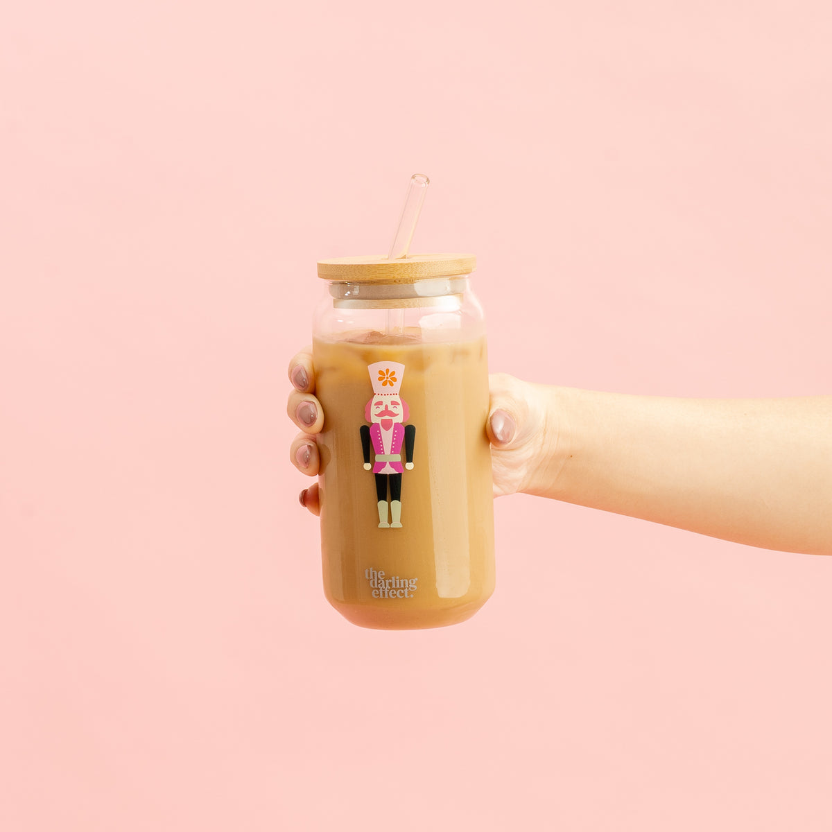 A hand holds The Darling Effect’s Round Glass Cup - Holiday Check, a glass cup of iced coffee with a bamboo lid and straw. The cup features a festive check pattern printed on it, set against a pink background.