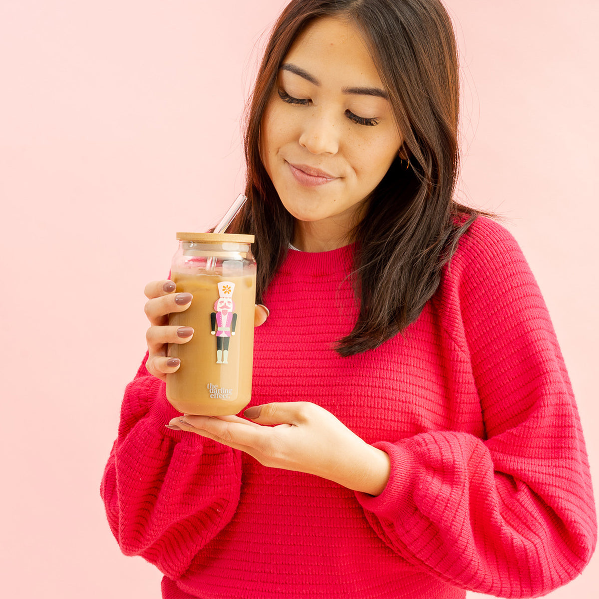 A woman in a bright pink sweater holds The Darling Effect’s Round Glass Cup - Holiday Check, filled with iced coffee and topped with a bamboo lid, featuring a festive nutcracker sticker and straw. She smiles while looking down at the drink, set against a pink background.